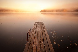Small wooden pier on still lake in autumnal foggy morning with rising sun