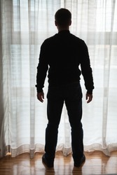 Young man stands near bright window with tulle and looks outside, back light silhouette photo