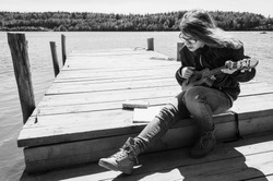 Caucasian teenager girl in glasses plays ukulele on a wooden pier, black and white outdoor photo