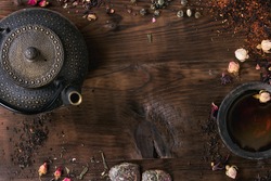 Food background with different black and green dry tea, rose buds cup of hot tea and iron teapot over dark wooden background. Tea drinking concept. Top view. Space for text.