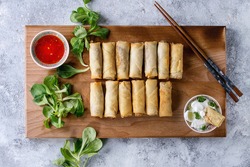 Fried spring rolls with red and white sauces, served on wood serving board with fresh green salad and wooden chopsticks over gray blue texture background. Flat lay, space. Asian food