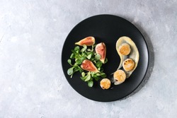 Fried scallops with lemon, figs, sauce and green salad served on black plate over gray texture background. Top view, copy space. Plating, fine dining