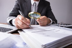 Close-up Of A Businessperson's Hand Looking At Receipts Through Magnifying Glass At Workplace