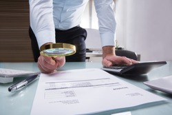 Close-up Of A Businessperson's Hand Holding Magnifying Glass Over Invoice At Workplace