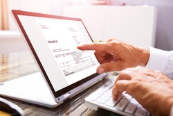 Close-up Of A Businessperson's Hand Analyzing Invoice On Laptop At Workplace