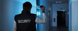 Rear View Of A Security Guard Standing In Corridor Of The Building Using Flashlight