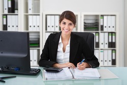 Portrait of confident female accountant writing on documents at desk in office