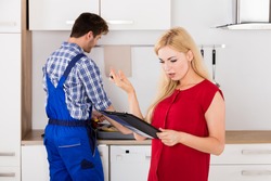 Young Frustrated Woman Looking At Expensive Bill On Clipboard With Plumber Standing In Kitchen