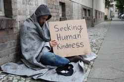 Male Beggar In Hood Showing Seeking Human Kindness Sign On Cardboard