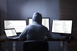 Rear View Of A Hacker Using Multiple Computers For Stealing Data On Desk