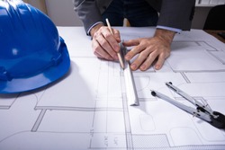 Close-up Of Architecture's Hand Working On Blueprint With Hardhat On Desk