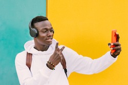 Young african American black man looking at smart phone while listening music with wireless headphones. Taking self portrait, selfie. Yellow and green wall wearing a white sweatshirt and a backpack