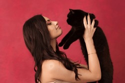 Beautiful woman holding a black cat on a pink background.