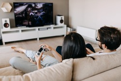 Back view of a couple of man and woman playing video game on a console, sitting on couch in living room.