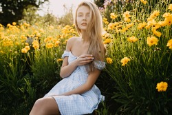 Young woman with long blonde hair, wearing country style dress, sitting in a garden with yellow flowers in golden hour.