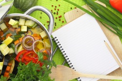 Blank recipe book with vegetable soup, kitchen equipment and veggies around them. Healthy cooking