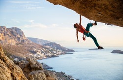 Caucasian man climbing challenging route going along ceiling in cave at sunset, against beautiful evening view