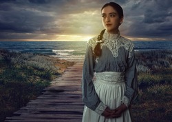 Young victorian girl in white dress and blue striped blouse on the coast at sunset.