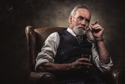 In chair sitting senior business man with cigar and whisky. Gray hair and beard wearing blue striped gilet and tie. Against brown wall.