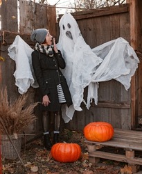 Boo. Vertical shot of scared young woman in dark clothes looking at creepy fake ghost with shocked face expression while standing at house backyard decorated with pumpkins during Halloween holiday