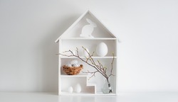 springtime Home minimalist interior with Easter decor. Branches with budding buds in a glass vase on shelves in the shape of a house, Easter eggs in a nest basket on a white background.