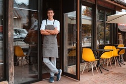 Portrait barista or waiter cafe or coffee shop owner against entrance, gesture inviting you to visit, smiling guy in apron standing outdoors being proud of his small local business