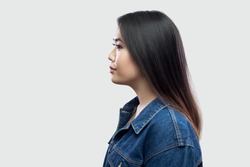 Profile side view portrait of calm serious beautiful brunette asian young woman in casual blue denim jacket with makeup standing and looking. indoor studio shot, isolated on light grey background.