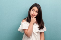 Portrait of thoughtful beautiful brunette young girl with black long straight hair in white dress standing, touching her chin, looking away and thinking. indoor studio shot isolated on blue background