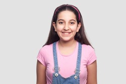 Portrait of happy beautiful brunette young girl in casual style, pink t-shirt and blue denim overalls standing and looking at camera with toothy smile. indoor studio shot, isolated on gray background.