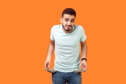 Portrait of frustrated worried brunette man with beard in casual white t-shirt turning out empty pockets showing I have no money gesture, bankrupt. indoor studio shot isolated on orange background