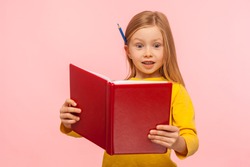 Shocking facts. Astonished smart little girl with pencil behind ear reading big book with surprised expression, amazed by story, learning encyclopedia. indoor studio shot isolated on pink background
