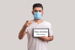 #StayAtHome. Man in protective mask holding Stay at home inscription and pointing to camera, warning of coronavirus quarantine, Covid-19 epidemic, preventive measures for spread of infectious disease.