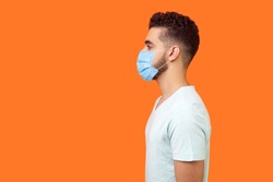 Side view of serious handsome brunette man with surgical medical mask standing with hands down and looking left, empty copy space for text. indoor studio shot isolated on orange background