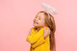 I love myself. Beautiful charming little girl with halo over head embracing herself and smiling from happiness, self-love concept, positive self-esteem. indoor studio shot isolated on pink background