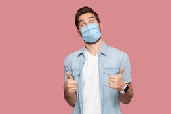 Portrait of young man with surgical medical mask in blue casual style shirt standing, thumbs up and looking at camera with surprised happy face. indoor studio shot, isolated on pink background.