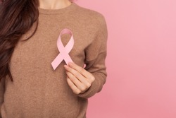Female health check awareness. Closeup of young woman in pullover holding pink ribbon, symbol of breast cancer, timely diagnosis, support of oncology patients. studio shot isolated on pink background