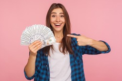 Portrait of joyful lucky girl in casual shirt happy to hold lot of money, pointing at dollar bills in hand and smiling excitedly, enjoying lottery win. indoor studio shot isolated on pink background
