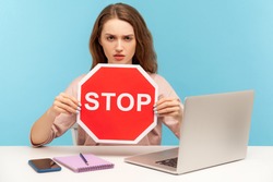Ban, forbidden access! Young woman holding stop symbol, warning with red traffic sign and looking angrily, sitting at workplace with laptop, home office. indoor studio shot isolated on blue background
