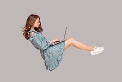 Hovering in air. Pretty girl ruffle dress levitating, typing keyboard using laptop for work online, surfing web social networks while flying in mid-air. indoor studio shot isolated on gray background