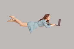 Hovering in air. Surprised excited girl ruffle dress levitating with laptop, typing keyboard, reading shocking news message on computer while flying in mid-air. studio shot isolated on gray background
