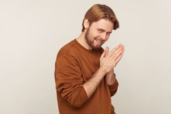 Portrait of sneaky sly scheming man with beard wearing sweatshirt rubbing palm as having cunning evil idea, devious plan in mind, thinking revenge. indoor studio shot isolated on gray background