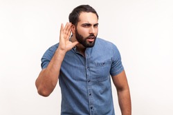 Concentrated nosy man with beard in blue shirt holding hand near ear, listening to interesting talks and private secrets, spying. Indoor studio shot isolated on white background