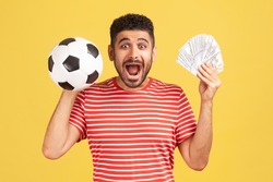 Extremely excited happy man with beard in striped t-shirt holding soccer ball and hundred dollar bills, screaming looking camera, betting and winning. Indoor studio shot isolated on yellow background