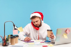 Man in santa claus hat sitting at workplace drinking coffee and talking landline telephone, writing down information into notepad, working on holidays. Indoor studio shot isolated on blue background