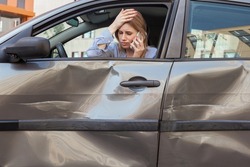 Blonde attractive woman with sad facial expression sitting behind wheel in crashed automobile and talking phone, keeping palm on her forehead, being in despair after road accident. Outdoor shot.