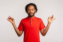 Portrait of relaxed bearded man with dreadlocks wearing red casual style T-shirt, standing with raised arms and doing yoga meditating exercise. Indoor studio shot isolated on gray background.