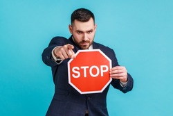 Hey you, access forbidden! Strict bossy man wearing dark official style suit, pointing to camera and holding Stop sign as symbol of prohibition. Indoor studio shot isolated on blue background.