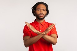 Portrait of young adult bearded man with dreadlocks wearing red T-shirt, crossing hands, gesturing warning or prohibition, meaning stop finish. Indoor studio shot isolated on gray background.