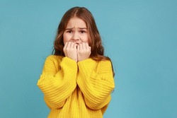Little girl biting nails with scared big eyes, feeling worried, anxious about childish mistake, wearing yellow casual style sweater. Indoor studio shot isolated on blue background.
