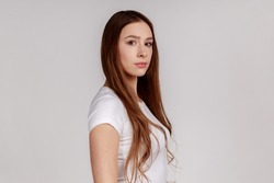Side view of portrait of strict bossy woman looking at camera, feels confident focused self-assured, expressing seriousness, , wearing white T-shirt. Indoor studio shot isolated on gray background.